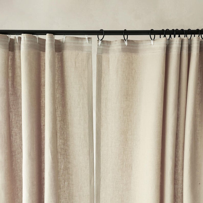Puede incluir: Cortinas de lino beige colgadas de una barra negra con anillas negras. Las cortinas son ligeramente transl&uacute;cidas, permitiendo el paso de algo de luz. La tela tiene un aspecto texturizado, con pliegues verticales.