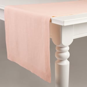 May include: A pale pink table runner made of linen. The runner is draped over a white table with a turned leg.