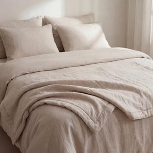 May include: A bed is made with a neutral-toned linen duvet cover, pillows, and a folded blanket. The bedding has a soft, textured appearance, creating a cozy and inviting atmosphere. The color palette is a warm, light beige.