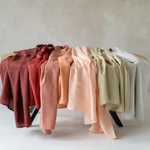 May include: A display of various colored linen fabrics draped over a curved surface. The colors range from deep red to light gray, including shades of orange, pink, and green. The fabrics are arranged to showcase their textures and hues.