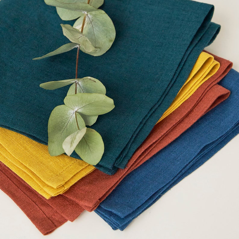 May include: A stack of four linen napkins in teal, mustard yellow, rust, and navy blue. The napkins are folded and arranged in a pile, with a sprig of eucalyptus leaves resting on top.