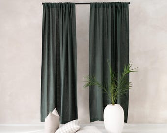 May include: Dark green linen curtains hanging from a black rod, with three neutral-toned pillows on the floor. A white vase holds a green plant, adding a touch of nature to the room. The curtains create a sense of privacy and elegance.
