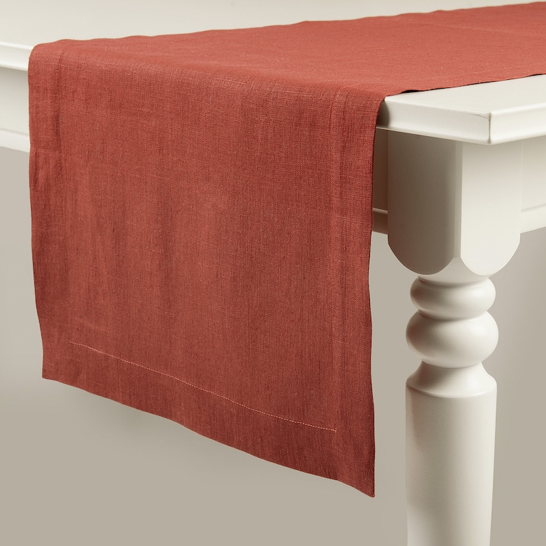 Puede incluir: Un camino de mesa de lino de color &oacute;xido con una textura sutil y un borde cosido.