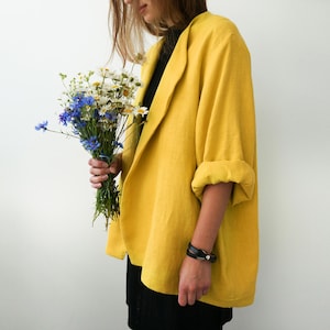 May include: A woman wearing a yellow linen blazer with a black top and black pants. She is holding a bouquet of white and blue flowers.