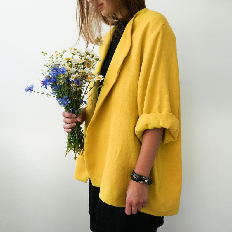 May include: A woman wearing a yellow linen blazer with a black top and black pants. She is holding a bouquet of white and blue flowers.