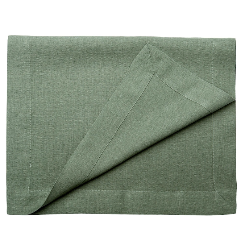May include: A sage green linen placemat with a folded corner, showing a detailed hem. The fabric has a textured appearance, suggesting a natural fibre. The placemat is rectangular and appears to be new.