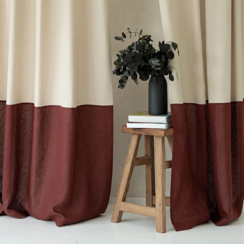 Puede incluir: Cortinas de bloques de color crema y burdeos. La parte superior de las cortinas es de color crema claro, mientras que la inferior es de un burdeos intenso. Las cortinas cuelgan en pliegues suaves. Un peque&ntilde;o taburete de madera sostiene un jarr&oacute;n negro con hojas verdes oscuras.