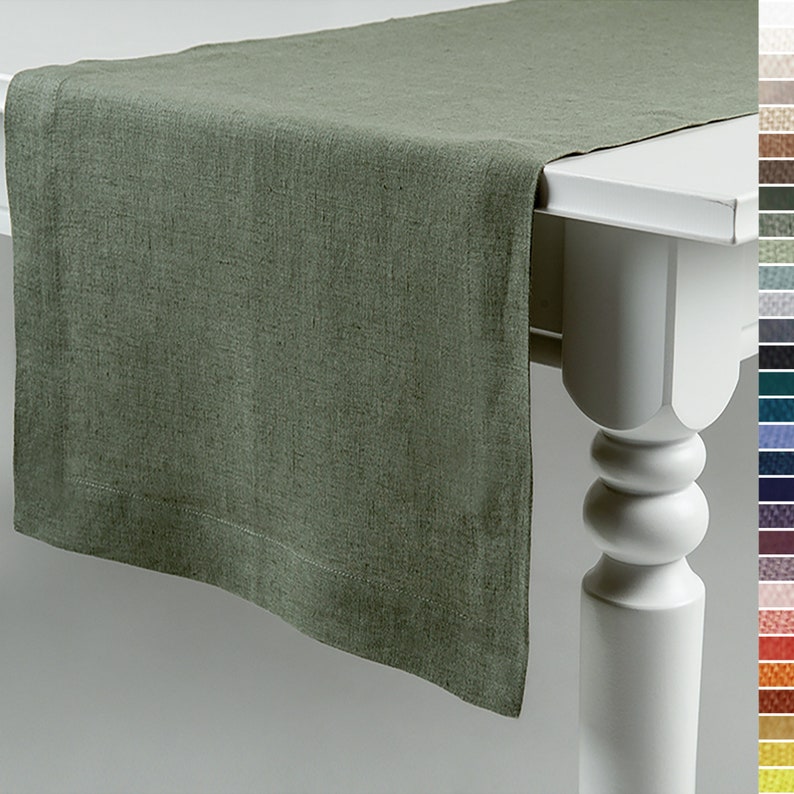 May include: A sage green linen table runner on a white table. The runner has a textured finish and a stitched hem. The table leg is visible on the right, with a colour palette alongside.