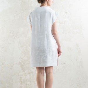 May include: A white linen dress with short sleeves and a straight silhouette. The dress is shown from the back, with the model standing on a white background.