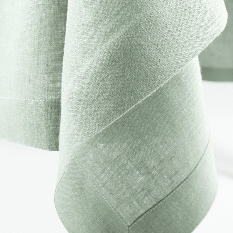 Peut inclure: Gros plan sur une nappe en lin vert sauge. Le tissu pr&eacute;sente un aspect textur&eacute; avec des d&eacute;tails de tissage visibles. Les bords sont soigneusement ourl&eacute;s avec une couture subtile de couleur plus claire, cr&eacute;ant un aspect &eacute;pur&eacute; et sur mesure. L'esth&eacute;tique g&eacute;n&eacute;rale est simple et &eacute;l&eacute;gante.