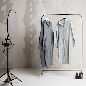 May include: Two linen shirts hanging on a black clothes rack. The shirt on the left is grey and the shirt on the right is light grey.