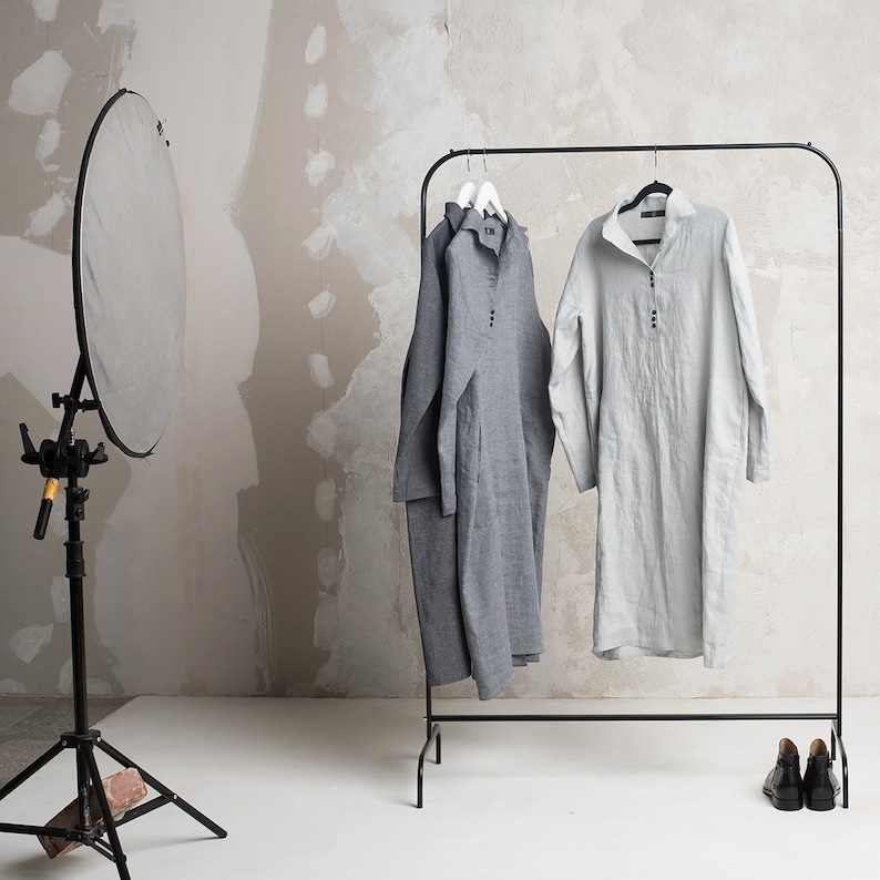 May include: Two linen shirts hanging on a black clothes rack. The shirt on the left is grey and the shirt on the right is light grey.