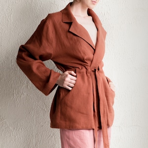 Può includere: Una giacca avvolgente in lino color ruggine con una silhouette lunga e fluida e una cintura a nodo. La giacca ha una profonda scollatura a V e tasche sui lati.