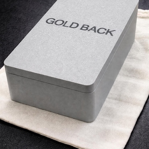 May include: A rectangular, light gray box with the text "GOLD BACK" printed on the top. The box is sitting on a white cloth, against a dark background. The box has a textured surface.