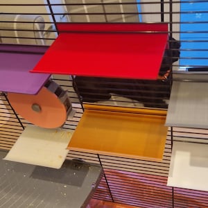May include: A multi-level pet cage with various colourful accessories. The cage features a purple, red, orange, yellow, grey, and white platforms. The cage is made of black wire and has a grey base.