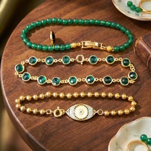 Puede incluir: Tres pulseras: una con cuentas verdes y cierre dorado, otra con piedras verdes facetadas redondas en engastes dorados, y una con cuentas doradas que presenta un dije central en forma de ojo con detalles en esmalte blanco, negro y azul claro.