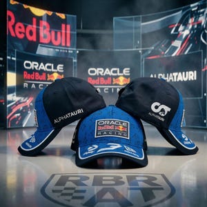 May include: Three baseball caps with the logos of Oracle Red Bull Racing and AlphaTauri. The caps are primarily black and blue, with white accents. The Red Bull logo is visible in the background.
