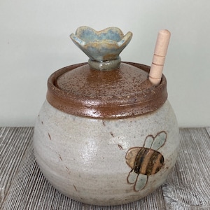 May include: Handmade ceramic honey pot with a flower-shaped lid handle and a wooden honey dipper. The pot is off-white with a brown rim and a painted bee design. A charming addition to any kitchen.