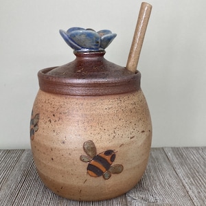 May include: A ceramic honey pot with a brown lid and a blue flower-shaped handle. The pot is decorated with painted bees and includes a wooden honey dipper. The pot has a speckled, tan finish.