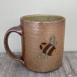 May include: A brown ceramic mug with a handle, featuring a hand-painted bee design. The bee has black and orange stripes with green wings. The mug has a speckled texture and a light green interior.