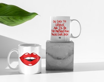 May include: Two white ceramic mugs. One mug features a red lip design. The other mug has the text "ON GOES THE LIPSTICK AND I'LL BE THE PRETTIEST MESS YOU'VE EVER SEEN" in red. The mugs are on a white surface.