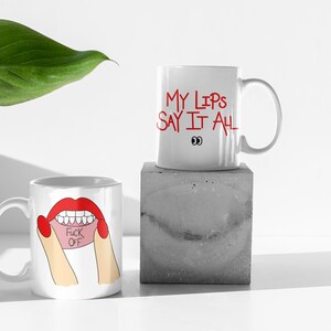 May include: Two white ceramic mugs with humorous designs. One mug features red lips with the words "FUCK OFF" on a pink tongue. The other mug has the text "MY LIPS SAY IT ALL" in red with cartoon eyes. The mugs are on a white surface.