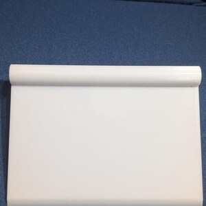 May include: A white, rectangular, plastic cutting board with a rolled edge. The cutting board is clean and appears unused. The background is a solid blue fabric.