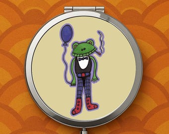 May include: A round, silver compact mirror featuring a cartoon frog in a tuxedo holding a blue balloon and a lit cigar. The frog is green with a black vest, plaid pants, and red boots. The background is a light yellow.