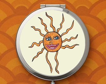May include: A round, silver compact mirror featuring a cartoon sun design. The sun has orange rays, a smiling face with blue eyes, and a cream-colored background. The mirror is set against an orange and brown patterned background.