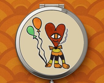 May include: A round, silver compact mirror with a cartoon heart character holding balloons. The heart has one eye, a bow tie, and is smoking. The background is a cream color.