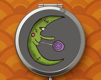 May include: A round, silver compact mirror with a cartoon crescent moon design. The green moon has stitches, a pink cheek, and is holding a purple lollipop against a gray background. The background is orange.