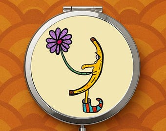 May include: A round, silver compact mirror featuring a whimsical cartoon banana character holding a purple flower. The banana is yellow with brown spots, wearing a colorful striped shoe. The background is a light yellow.