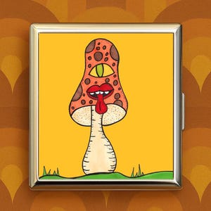 May include: A gold-colored metal case with a vibrant, cartoonish mushroom illustration. The mushroom has a spotted orange cap, a single green eye, and a red tongue. The background is yellow with a green base and grass.
