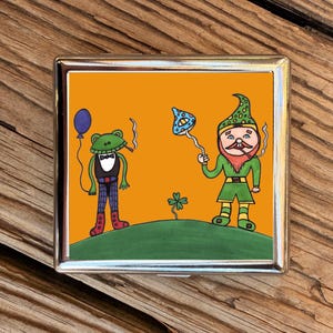 May include: A silver cigarette case with a colourful illustration. The artwork shows a frog in a bow tie and a gnome, both smoking. A blue balloon and a mushroom-shaped object are also depicted, along with a four-leaf clover.