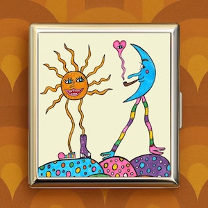 May include: A silver metal cigarette case featuring a whimsical illustration. The artwork depicts a smiling sun with long legs and a blue moon figure smoking a pipe, both walking on colorful polka-dotted hills. The background is a light cream color.