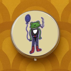 May include: A circular pill box with a silver-coloured frame, depicting a cartoon frog in a tuxedo, holding a blue balloon and a lit cigar. The frog is green with a black waistcoat, bow tie, and plaid trousers. The background is pale yellow.
