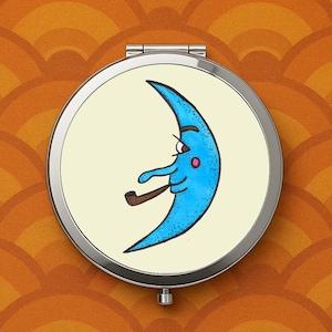 May include: A round, silver compact mirror featuring a cartoon crescent moon with a blue face, smoking a pipe. The moon has a pink cheek and is set against a cream-colored background. The mirror is on a patterned orange background.