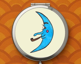 May include: A round, silver compact mirror featuring a cartoon crescent moon with a blue face, smoking a pipe. The moon has a pink cheek and is set against a cream-colored background. The mirror is on a patterned orange background.