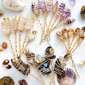 May include: A collection of crystal hair pins, each with gold wire detailing. The crystals are in shades of clear, purple, and brown. Seashells and small stones are arranged around the pins on a white background.