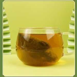 May include: A clear glass mug filled with amber-colored tea, with a tea bag steeping inside. The mug has a rounded shape and a handle. The background is a light green color, with a decorative paper fan on the left side.