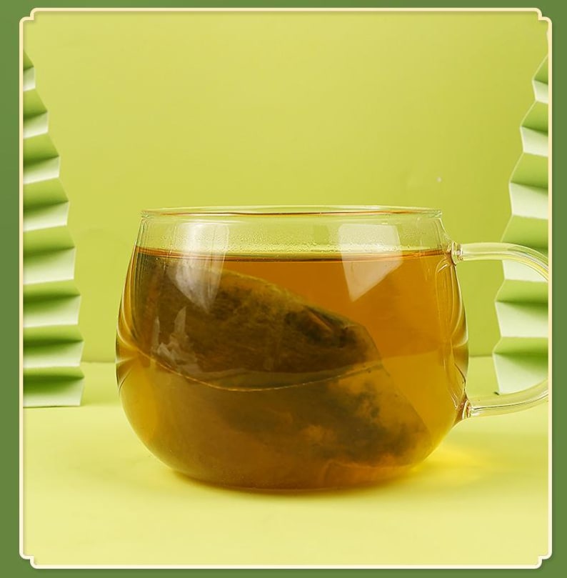 May include: A clear glass mug filled with amber-colored tea, with a tea bag steeping inside. The mug has a rounded shape and a handle. The background is a light green color, with a decorative paper fan on the left side.