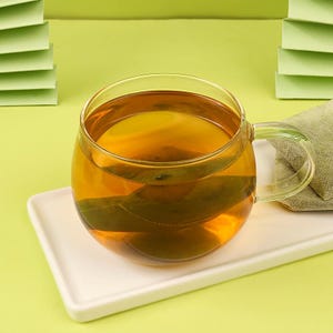 May include: A clear glass teacup filled with amber-colored tea, resting on a white rectangular plate. A tea bag is visible inside the cup. The background is a light green color.
