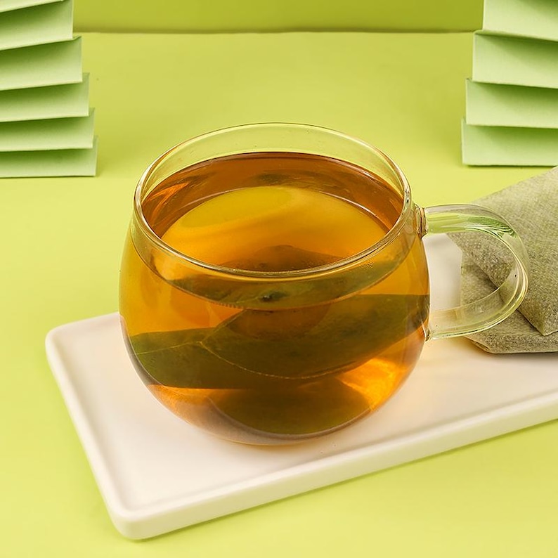 May include: A clear glass teacup filled with amber-colored tea, resting on a white rectangular plate. A tea bag is visible inside the cup. The background is a light green color.