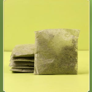 May include: A stack of five square, olive-green tea bags filled with loose tea leaves. The tea bags are made of a textured, semi-transparent material, and the tea leaves are visible inside. The background is a solid, light green.