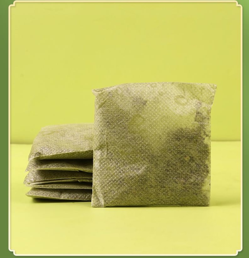 May include: A stack of five square, olive-green tea bags filled with loose tea leaves. The tea bags are made of a textured, semi-transparent material, and the tea leaves are visible inside. The background is a solid, light green.