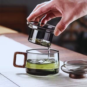 May include: A clear glass teacup with a wooden handle is being filled with tea from a glass infuser. The tea is a light green color. The infuser is held by a hand. A second glass lid with a wooden base sits nearby.