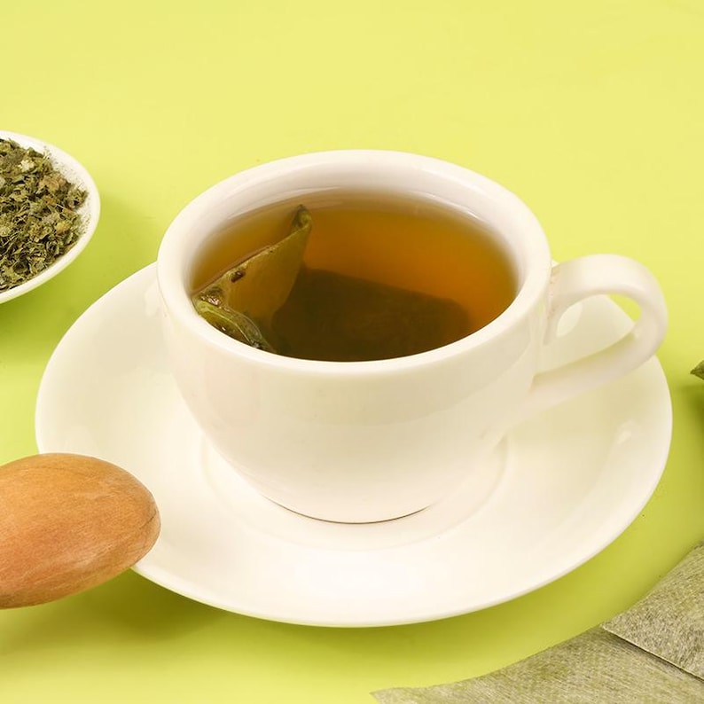 May include: A white teacup and saucer filled with tea, with a tea bag visible. A small white bowl of dried tea leaves is to the left. The background is a light green color. A wooden spoon is in the lower left corner.