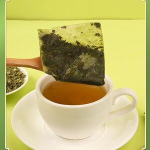 May include: A white teacup and saucer filled with tea, with a tea bag held above it on a wooden spoon. The tea bag is square and filled with green tea leaves. A small bowl of loose tea leaves is in the background.