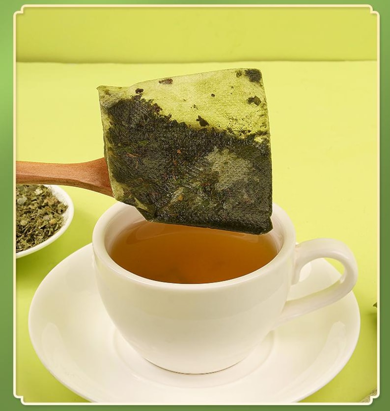 May include: A white teacup and saucer filled with tea, with a tea bag held above it on a wooden spoon. The tea bag is square and filled with green tea leaves. A small bowl of loose tea leaves is in the background.