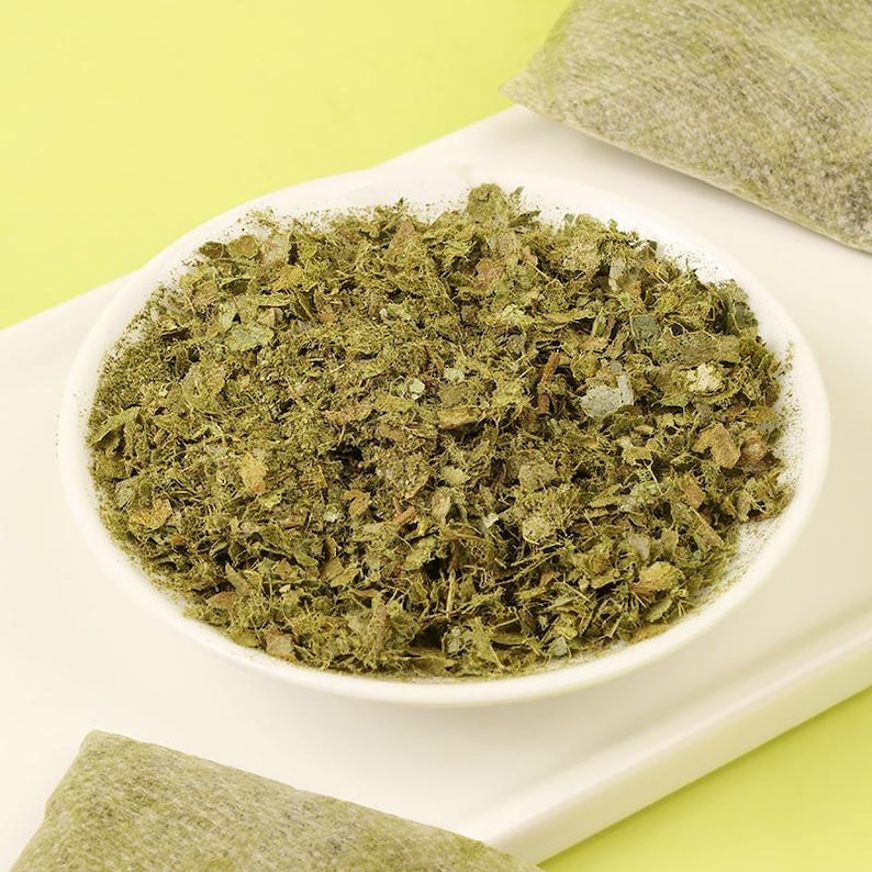 May include: A white bowl filled with dried, chopped green herbs, possibly for culinary or medicinal use. The herbs have a textured appearance and varying shades of green. A green package is visible in the background, suggesting a packaged product.